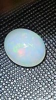 Close-up photo showing a smooth oval cabochon opal stone with multicolored flashes on a textured black background.