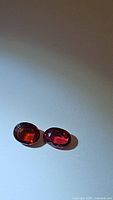 Pair of deep red purple rubelite and fired red orange untreated tourmaline gemstones photographed on a plain light background showing their oval cut and faceted surfaces.