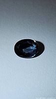 Oval shaped deep London blue topaz gemstone lying flat on white surface under light showing facets and color.