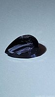 Photo showing a deep purple, pear-shaped iolite gemstone on a white background with light reflections highlighting facets.