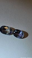Two oval-cut purple tanzanite gemstones on a plain background showing facets and color under direct light.