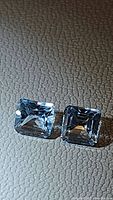 Two natural light sky blue topaz gemstones, princess cut, placed on a textured dark surface showing their square shape and clarity.