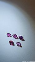 Five marquis cut purple-red rubies with visible faceting, shown scattered on a white surface with light reflection.