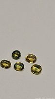 Five faceted oval sapphires laid out on a white background showing yellow, green, and blue-green tones.