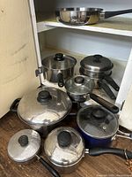 Photo of assorted pots and pans with lids, one with steamer insert, stored in a kitchen cabinet and on the floor