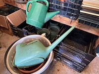 Green plastic watering cans and some plastic planting pots inside buckets and crates on a shelf.