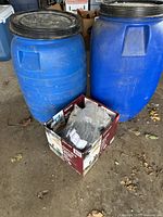 Photo showing two large blue plastic barrels and a cardboard box with bird seeds inside on a garage floor.