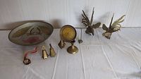 Lot of brass and copper decorative pieces arranged on a white surface in front of white panel walls. Includes a round painted metal bowl, fighting cock figurines, small bells, candle stick, incense burner, and mouse ring holder.