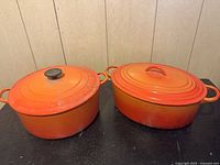 Photo showing two orange cast iron casserole dishes with lids, round on left and oval on right, placed on a black surface against wood panel background.