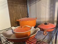 Four pieces of orange enameled cast iron cookware including frypan with lid, large pot with lid, small pot with lid, and oval casserole dish arranged on wire rack.