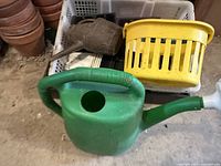 Green plastic watering can in front with a yellow plastic basket and brown watering can behind in white plastic bin, with terracotta pots stacked on left