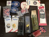Photo showing various sets of darts, new and used dart flights, and dart league patches arranged on a table.