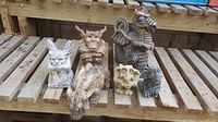 Wide view of six garden statues: one dragon and five gargoyles arranged on wooden slats in a greenhouse shelf.