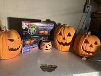 Three plastic carved pumpkins, orange with green stems, and two boxed strands of 10 pumpkin string lights, plus small ceramic pumpkin candle holder on table against basement wall.