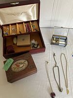Two wooden jewelry boxes, one open showing earrings, pins, and brooches, alongside a smaller wooden box with bird artwork, three pendants, and a small glass music box with etched lid.