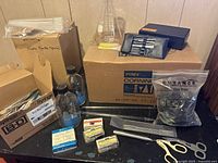 Boxed pyrex beakers, plastic pipettes box, glass bottles, and micro slides shown atop a black table, stainless steel tools pouch and black pH meter hard case visible