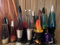 Photo of nine lava lamps of different colors, sizes, and base types grouped together against wood paneling background.