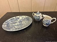 Grouped photo of Myott The Hunter 13-inch serving platter, sugar bowl with lid, and creamer on a dark surface against wooden panel background.