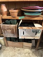 Shelf containing unglazed terracotta clay pots stacked on the top left, assorted colored round plastic trays stacked on the top right, and cardboard boxes below filled with green plastic square pots.