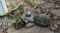 Lot showing two frog statues and one engraved rock statue on outdoor ground with leaves, stone chips and wooden edge visible