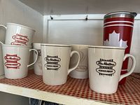 Seven Tim Hortons mugs on shelf; four ceramic and two plastic mugs with varying logos plus one red and white travel tumbler.