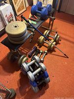 Weight bench on red carpet with a pile of York barbell plates stacked on it, several dumbbells and hand weights below, an extendable walking stick resting on bench, and a hand grip exerciser.