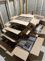 Multiple cardboard boxes filled with rows of jewel-case audio CDs stacked on a concrete floor.