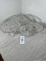 Three clear antique pressed glass cake stands with distinct patterns and pedestal bases shown together on beige cloth surface.