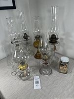 Six pressed glass oil lamps of varying sizes with chimneys on a table, accompanied by a vintage jar of wicks.