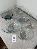 Six Pyrex glass mixing bowls of varying sizes and styles arranged on a gray cloth surface showing clear, ribbed, etched, blue, and smoky glass bowls.