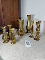 Group of 8 vintage brass vases displayed on a cloth surface with a wood background, showing varying shapes and sizes, including decorative and plain finishes.