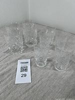 Group of 9 cut crystal tumblers on beige cloth, 4 larger in back, 5 smaller in front, all clear patterned with one large tumbler chipped at rim.