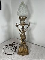 Full view of Art Deco lamp with a gilded spelter female figure base holding a horizontal bar supporting a frosted flame glass shade, power cord visible on surface.