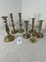 Lot of seven vintage solid brass candlesticks arranged on fabric surface, showing range of sizes and styles.