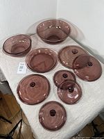 Full lot of cranberry Pyrex glassware including mixing bowl, casserole, pie plate, and lids arranged on a beige cloth surface.