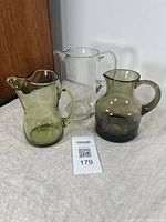 Photo showing all three vintage blown glass pitchers grouped together on a cloth surface against a white and wooden background.