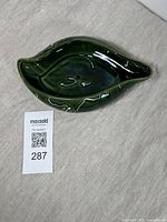Top and angled side view of green glazed leaf-shaped ceramic dish on neutral fabric surface with a lot tag.