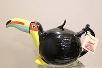 Side view of glossy black toucan teapot with colorful beak and yellow feet, displaying full bird shape and body detail.