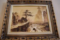 Front view of the framed Asian landscape painting showing mountains, trees, bridge and river in earthy tones.