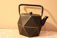 Side angle of cast iron teapot showing geometric polygonal design, black color with gold outline accents, handle and spout.