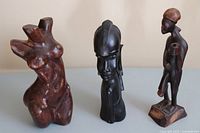 Photo showing all three wooden sculptures together: an abstract female torso, African head bust, and standing male figure with stick.