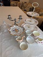Full view of assorted vintage fine china and servingware on table including three-tiered stand, scalloped plate, creamers, candle holder and covered glass dish.