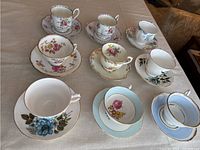 Nine vintage fine china teacup and saucer sets arranged on a table with a beige tablecloth. Various floral designs and some blue and gold accents visible.