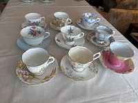 Full view of all nine teacup and saucer sets arranged in rows on white tablecloth showing variety of designs and colors.