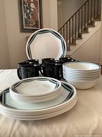 Overview of dinnerware set showing stacked dinner plates, salad plates, bowls, and black mugs arranged on table.