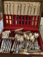 Flatware collection shown in original wood box with red felt interior, includes knives, forks, spoons, and serving pieces with ornate handle designs.