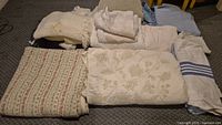 Full view of assorted folded bedding and linens including quilted bedspreads, sheets, and a hand embroidered baby blanket