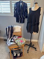 View of multiple women's clothing items including a black sleeveless layered top on a dress form, a blue and white wrap-style top hanging on a wall, and several folded tops on a chair displaying variety.