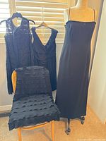 Photo showing four black dresses displayed on a dress form and a chair near window blinds, highlighting various styles including lace, layered fabric, and beaded details.