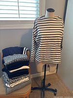 Photo displaying a stack of folded knitwear and activewear pieces on a chair with a white and navy striped top displayed on a dress form. The items show variety of colors including navy, grey, white and patterns including stripes and polka dots.